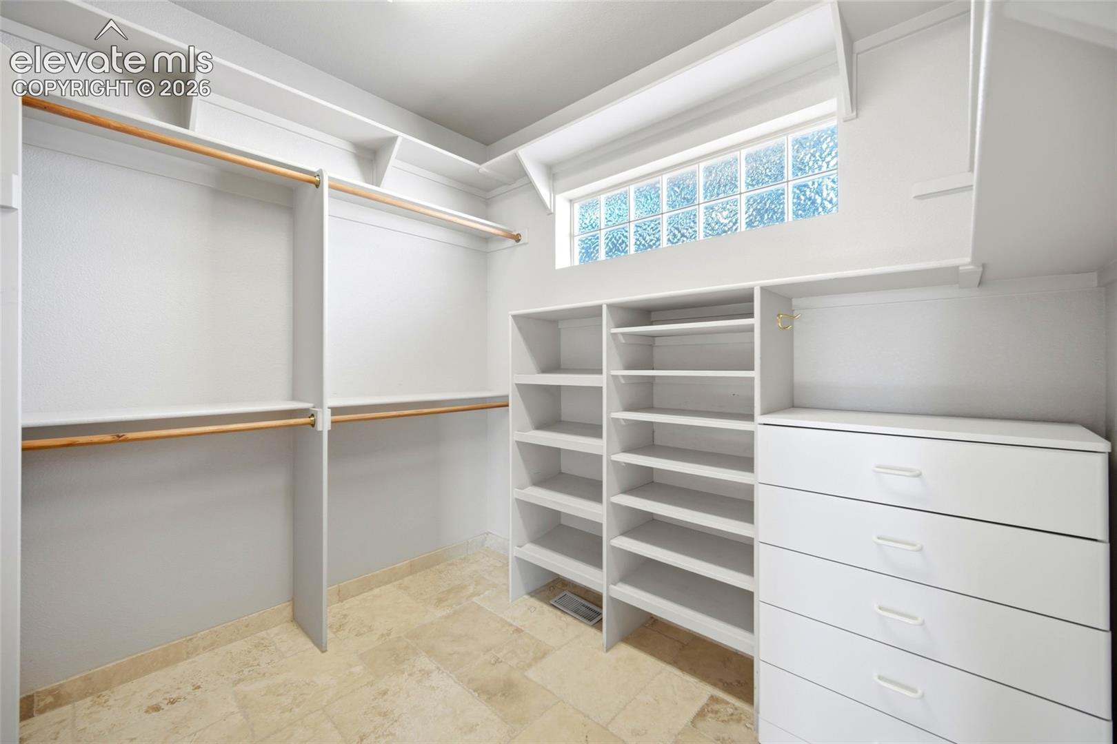 Walk-in closet featuring stone-finish tile flooring, white built-in shelving, integrated storage drawers, wooden hanging rods, and a textured glass transom window