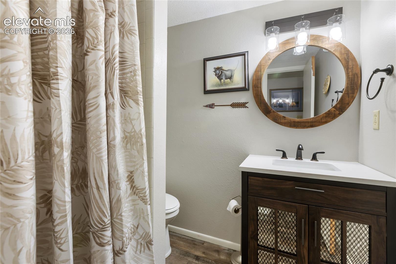 Wood-finish flooring, a dark wood-finish vanity with mesh cabinet inserts, a white countertop with an integrated sink, and a circular wood-framed mirror