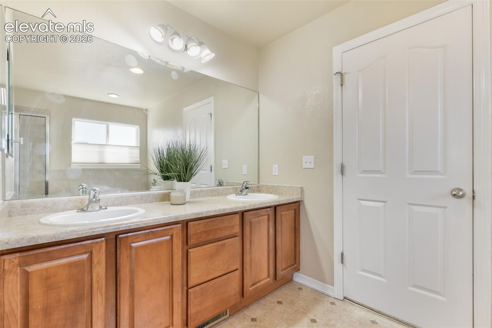 Dual undermount sinks with chrome faucets, wood-finish cabinetry, a large wall-mounted mirror, and overhead vanity lighting