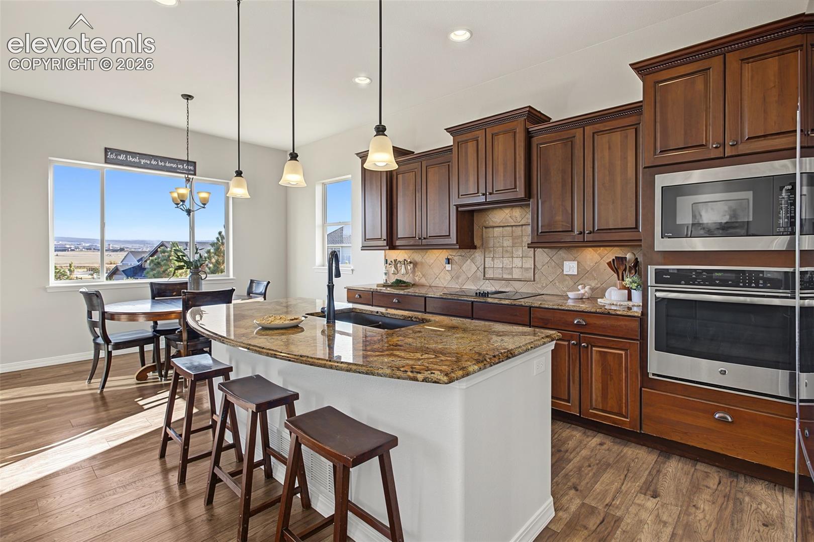 Kitchen featuring rich wood cabinetry, stone countertops, integrated stainless steel double wall ovens, wood-finish flooring, and a large center island with a drop-in sink and pendant lighting