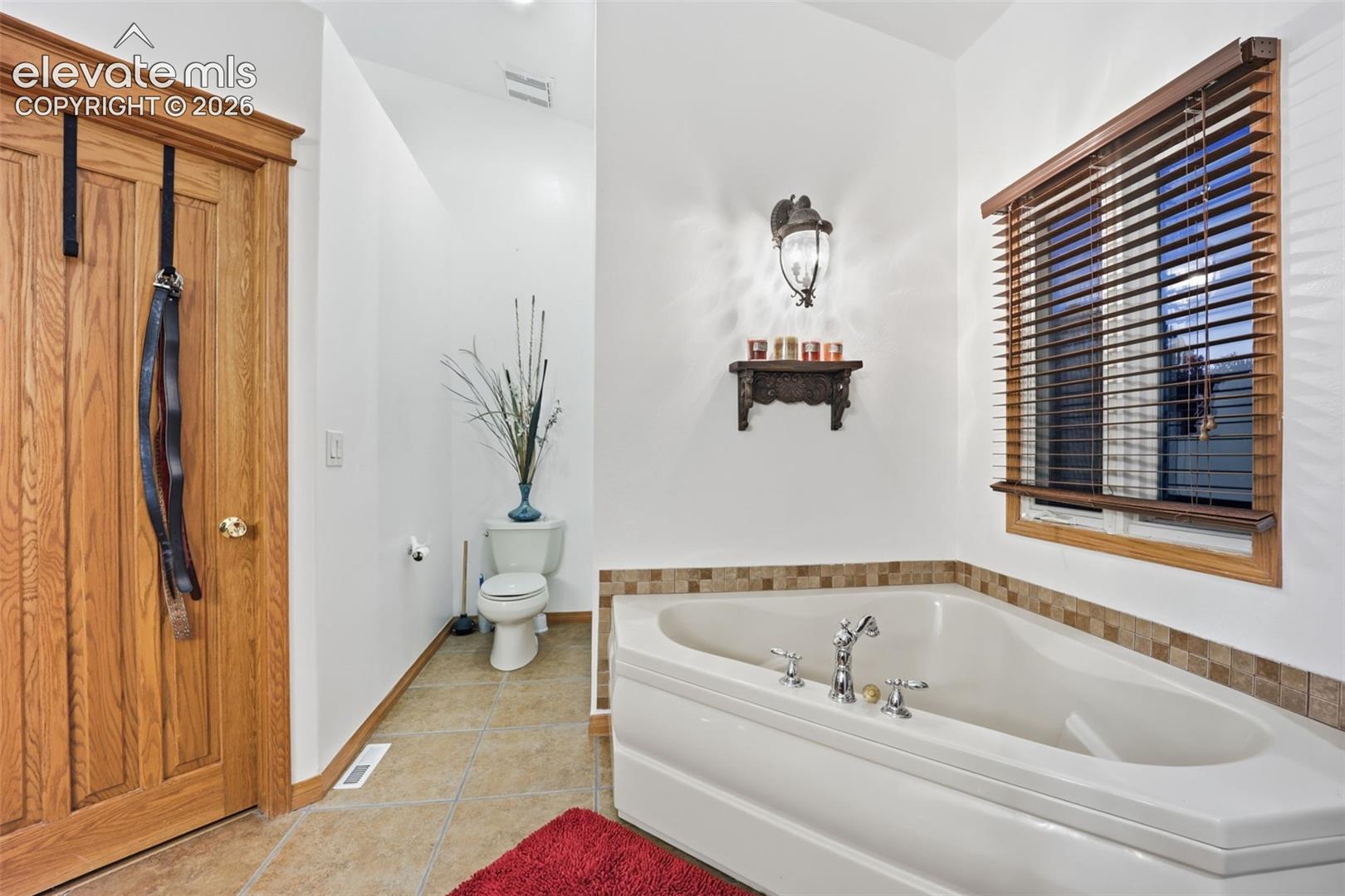 Corner soaking tub with polished chrome fixtures, tile surround, wood-slat window blinds, and overhead wall sconce