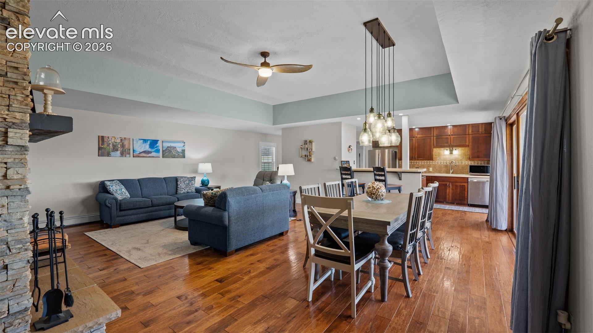 Open-concept living space featuring wood-finish flooring, a tray ceiling, a multi-light pendant chandelier, a stone-accented fireplace, and kitchen cabinetry with a breakfast bar