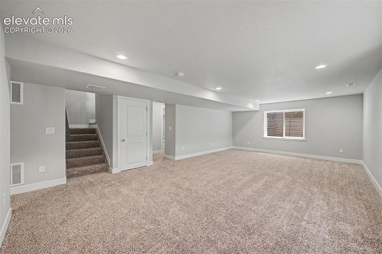 Spacious carpeted room featuring recessed lighting, a window with horizontal blinds, a white two-panel door, and a carpeted staircase