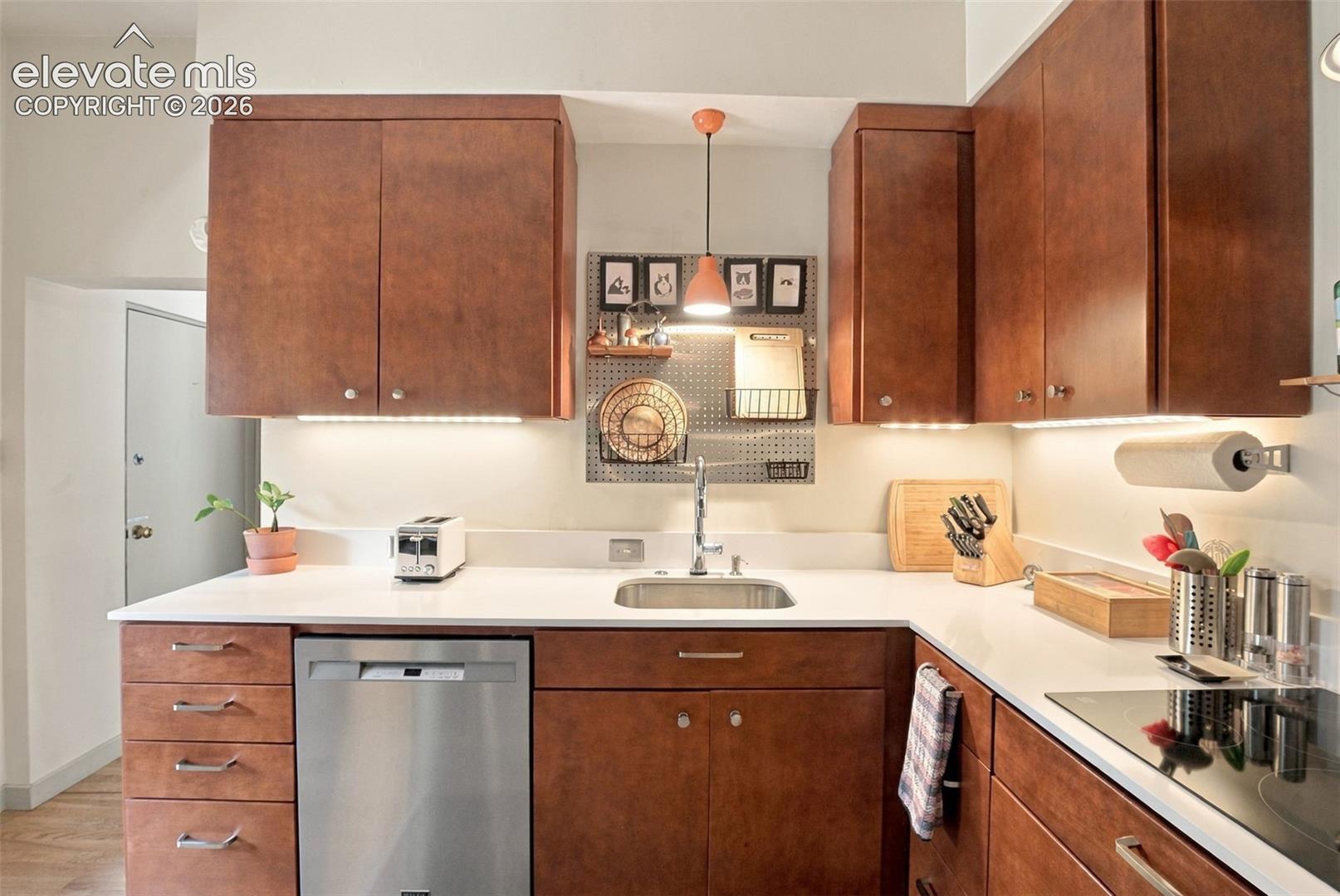 Kitchen featuring wood-finish cabinetry, solid surface countertops, stainless steel undermount sink, under-cabinet lighting, and built-in stainless steel dishwasher