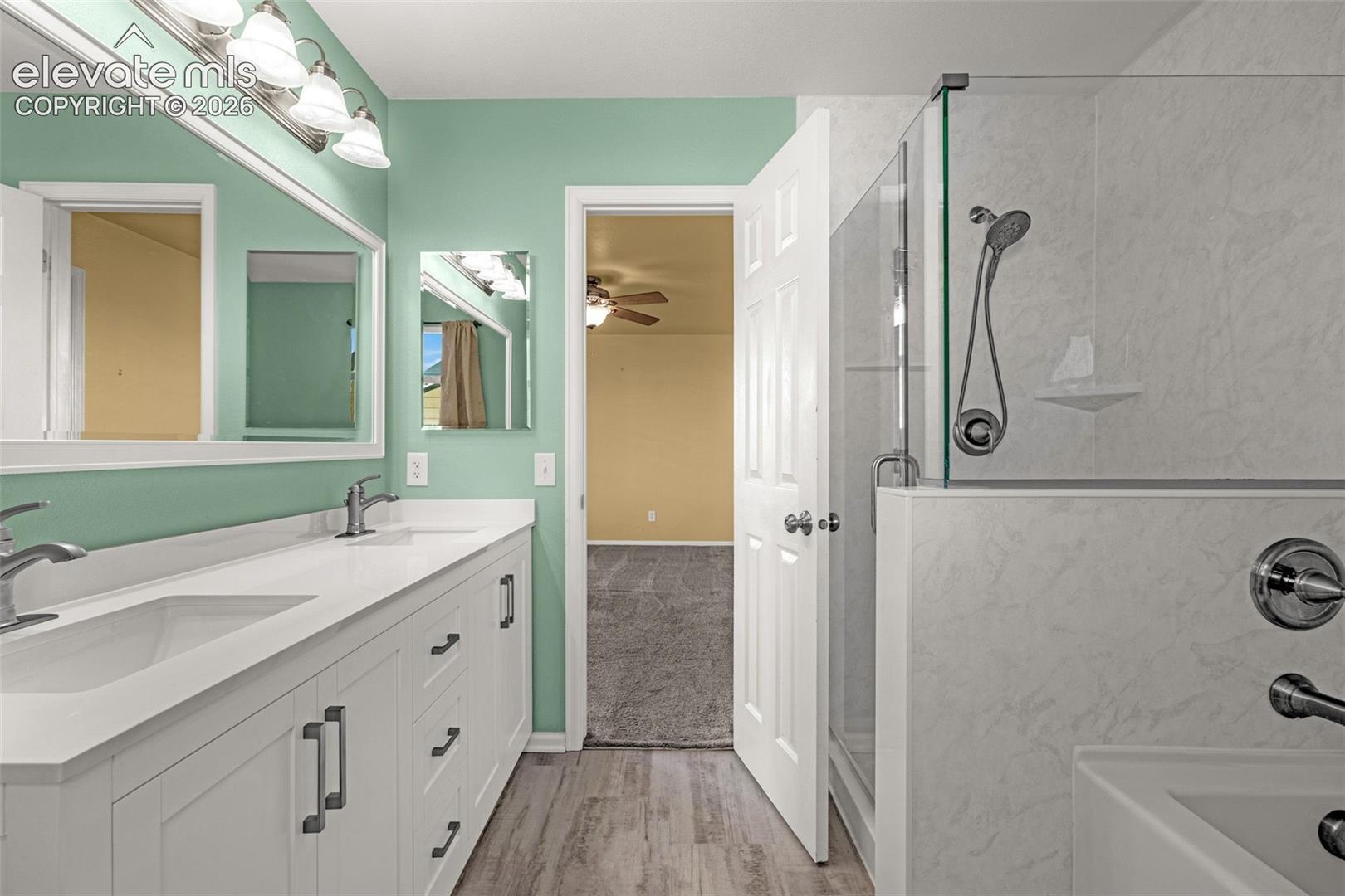 Dual-sink vanity with white cabinetry and quartz countertop, walk-in glass shower enclosure, integrated shower wall surrounds, and wood-finish flooring