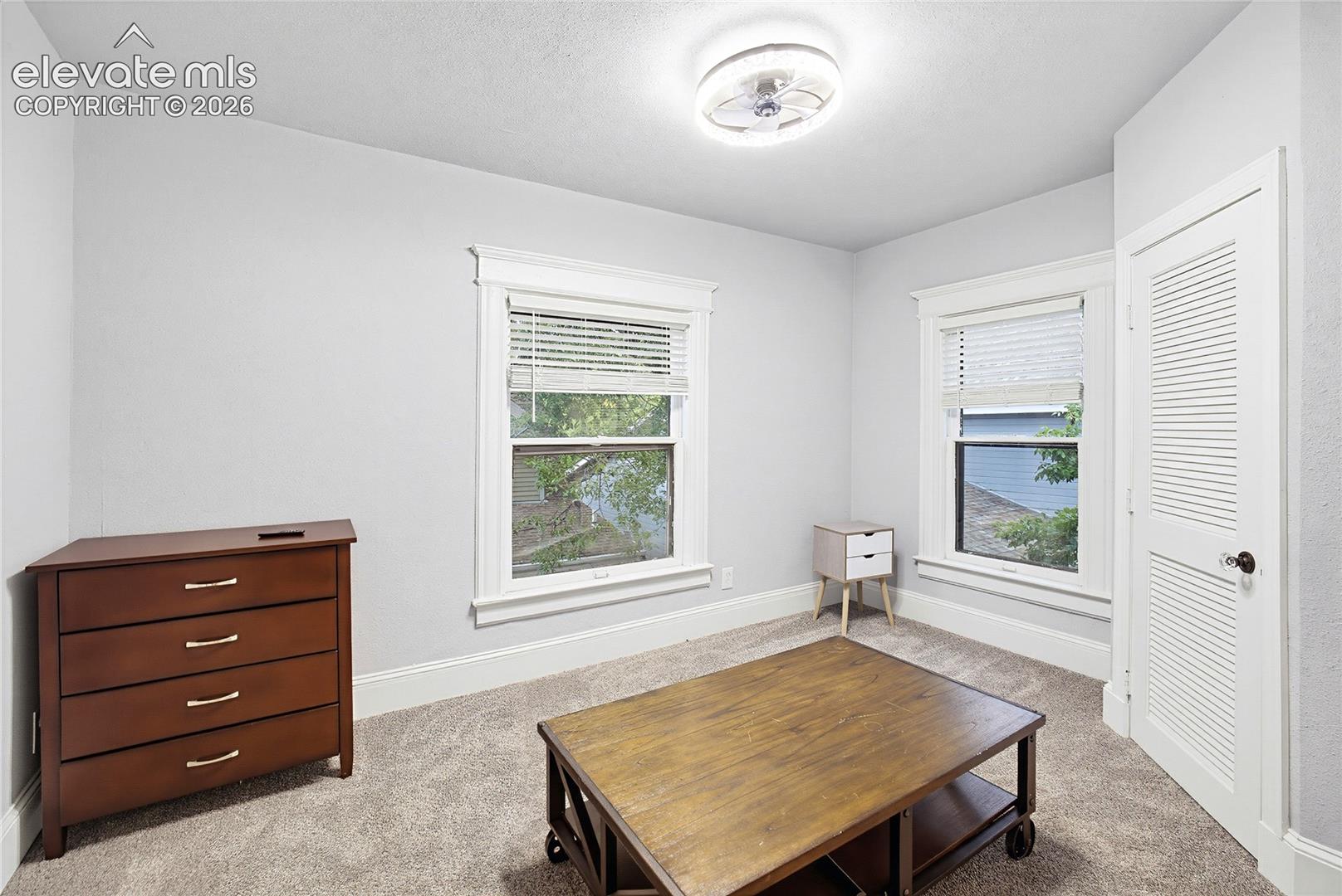 Neutral wall finishes, carpeted flooring, two windows with blinds, ceiling fan, and a louvered door