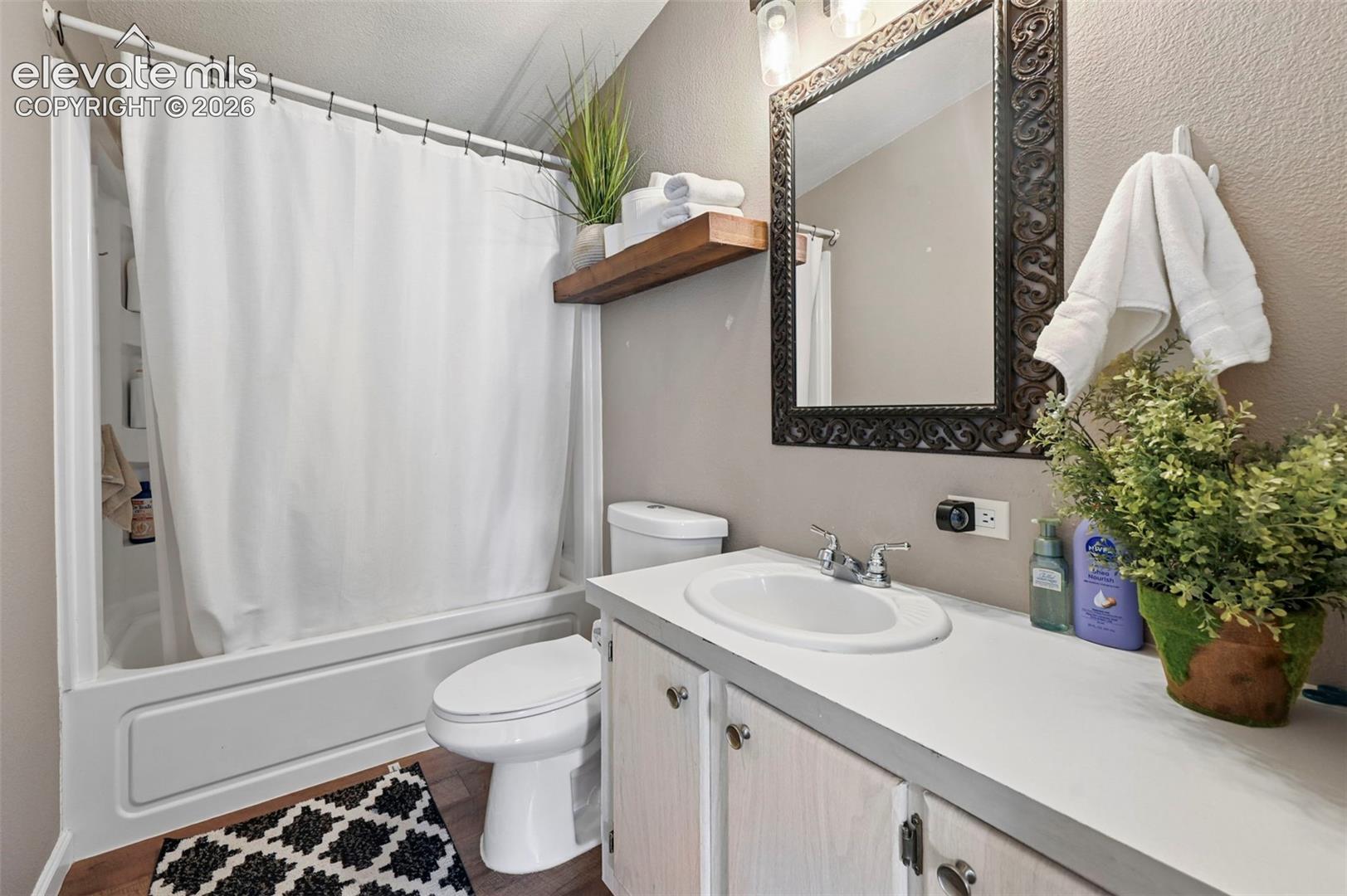Bathroom featuring a white vanity cabinet, solid-surface countertop with an integrated basin, wood-finish flooring, a wall-mounted wood shelf, and a framed mirror