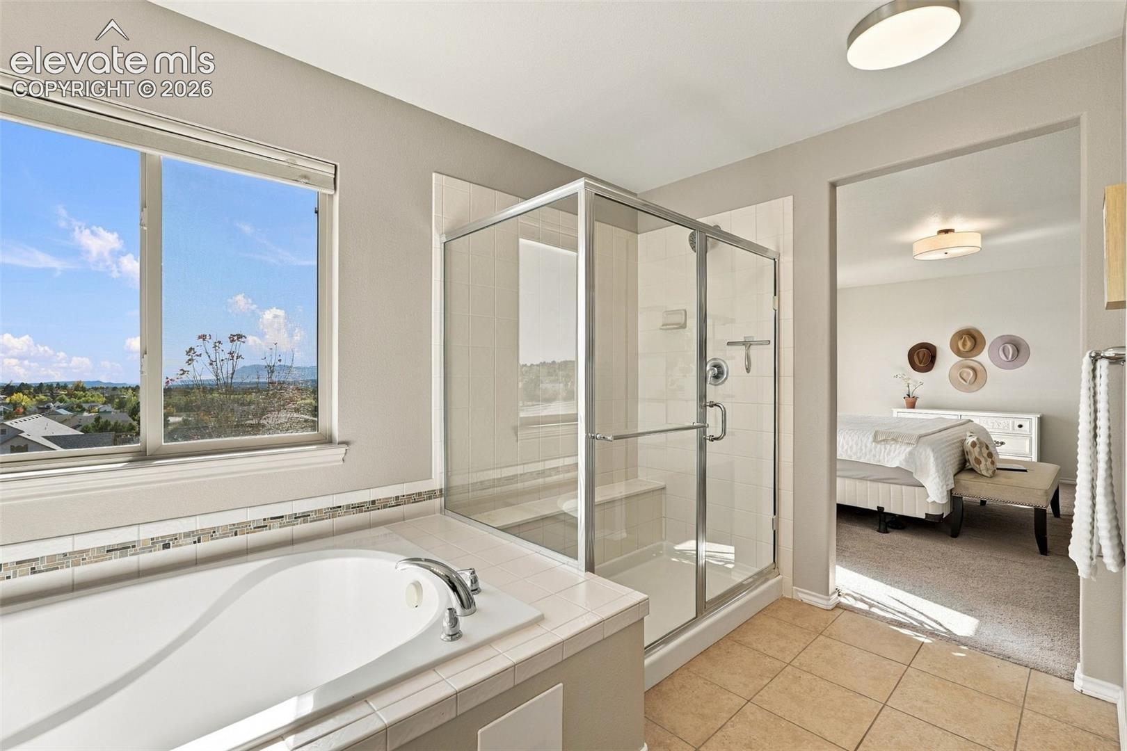 Deep soaking tub with tile surround, glass-enclosed shower featuring a built-in bench, large picture window with landscape scenery, and tile flooring