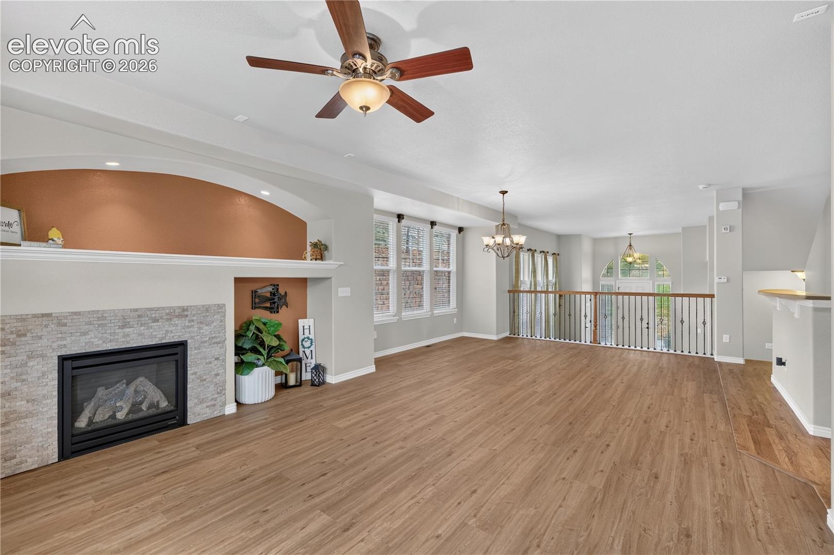 Fireplace with stone-style surround, arched recessed alcove, wood-finish flooring, ceiling fan with integrated lighting, and decorative metal baluster railing