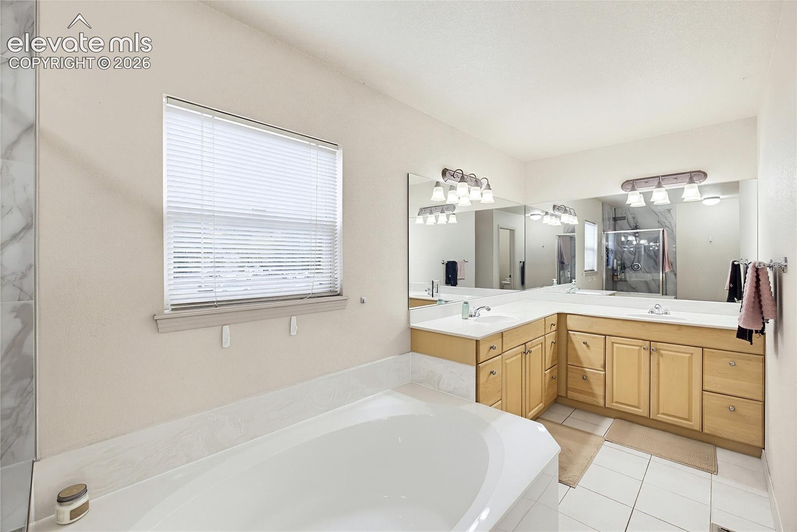 Built-in corner soaking tub, dual vanity with undermount sinks, extensive vanity mirror, light wood-finish cabinetry, and tiled flooring