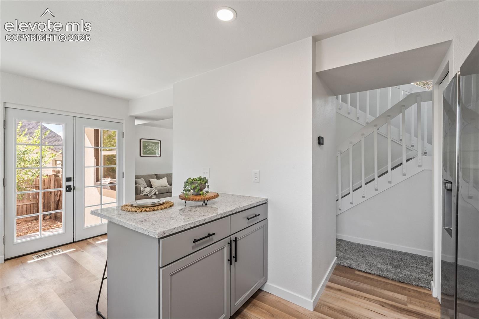 Kitchen island with stone-finish countertop and shaker-style cabinetry, wood-finish flooring, glass-paned double doors, and staircase with painted railing