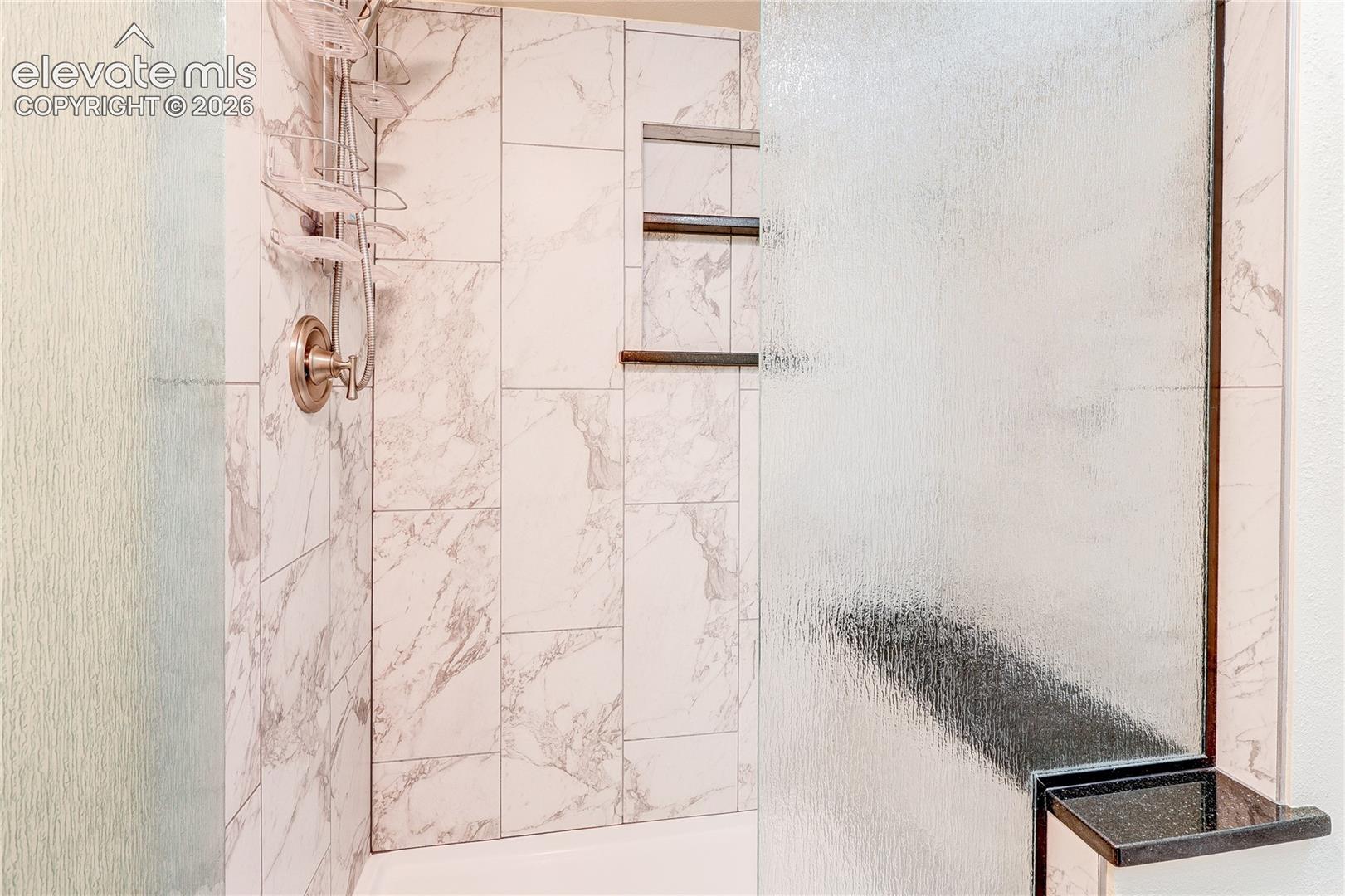 Shower enclosure featuring vertical veined tile wall surround, built-in recessed shelving, a brushed metal shower fixture, and a textured glass panel with a black-trimmed corner shelf