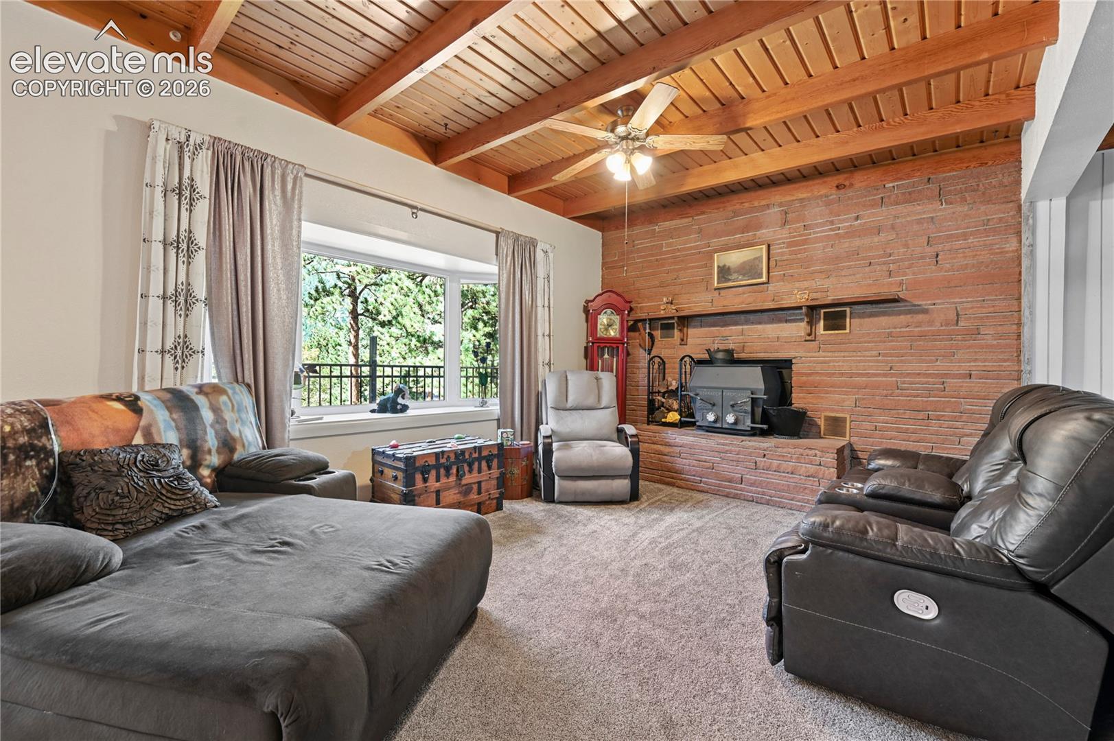 Exposed wood beam ceiling, brick accent wall with fireplace, bay window, and ceiling fan