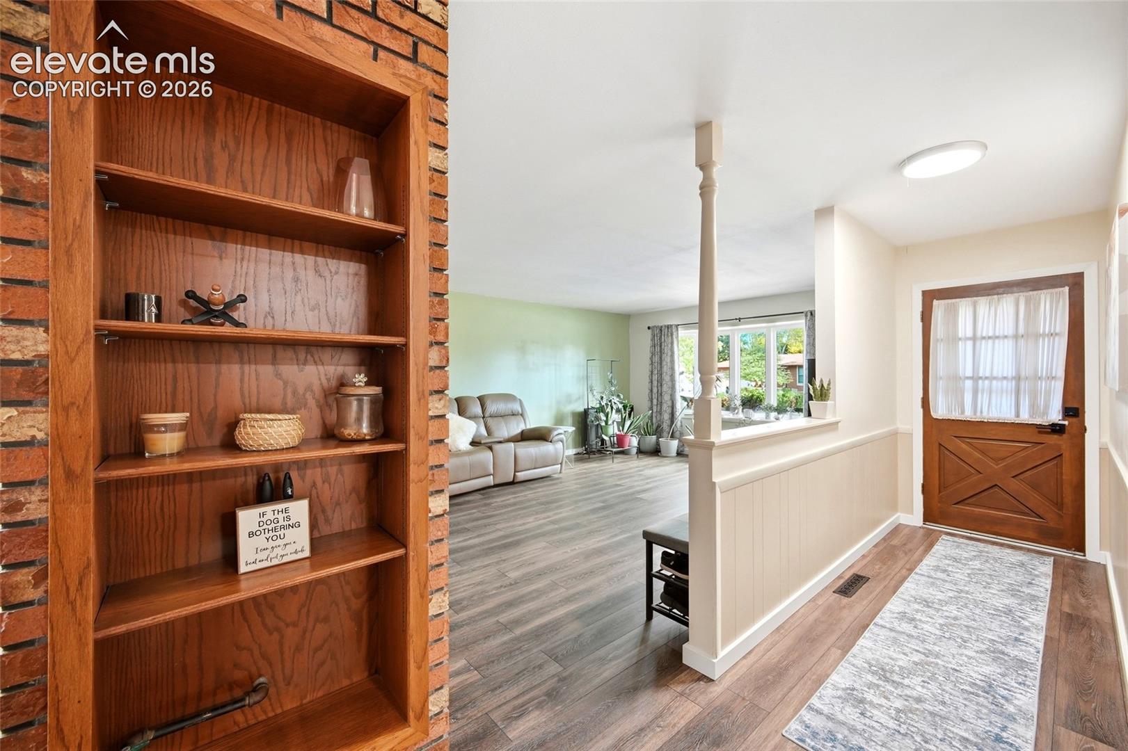 Entryway featuring a wood-finish door with an integrated window, built-in wooden shelving recessed into a brick accent wall, wood-finish flooring, and a half-wall partition with a column support