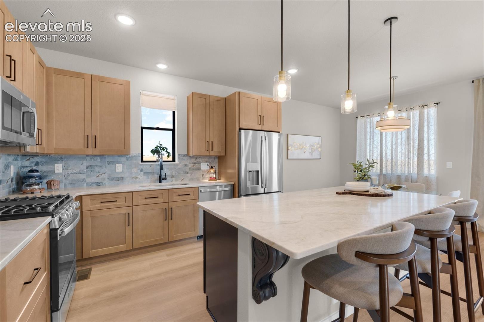 Kitchen featuring light wood-finish cabinetry, stainless steel appliances, a light-toned countertop island, a tiled backsplash, and recessed and pendant lighting