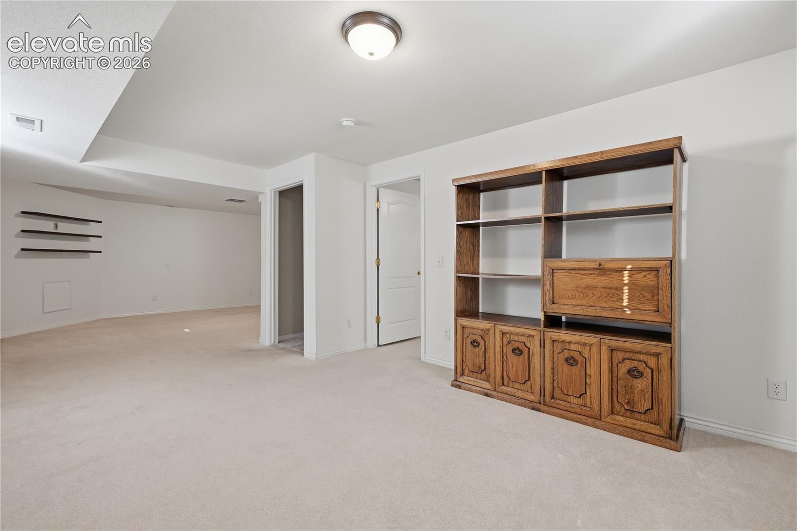 Neutral interior featuring built-in wooden cabinetry, wall-mounted shelving, carpeted flooring, and a flush-mount ceiling light fixture