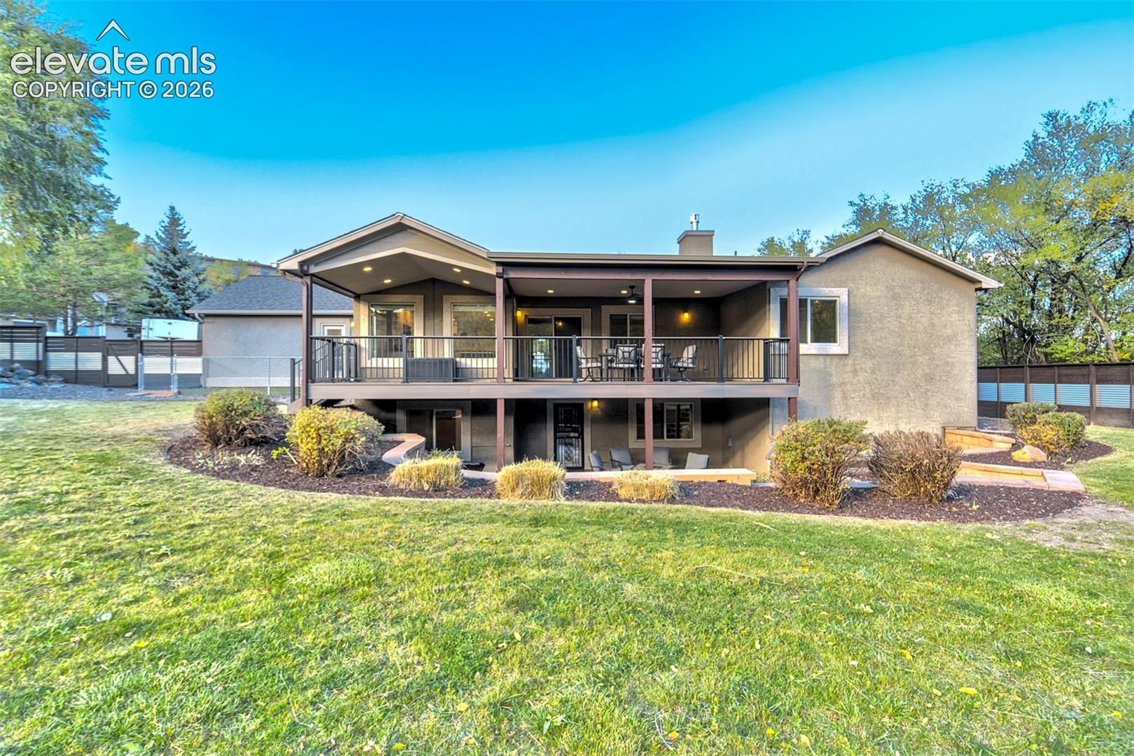 Stucco exterior residence featuring an expansive elevated deck with metal railing, covered patio space, and landscaped garden beds