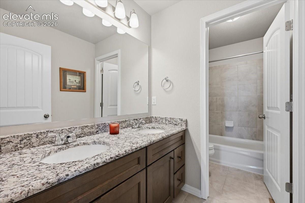 Dual-sink vanity with stone-finish countertop, dark wood cabinetry, large frameless wall-mounted mirror, and tile-surround shower-tub combination