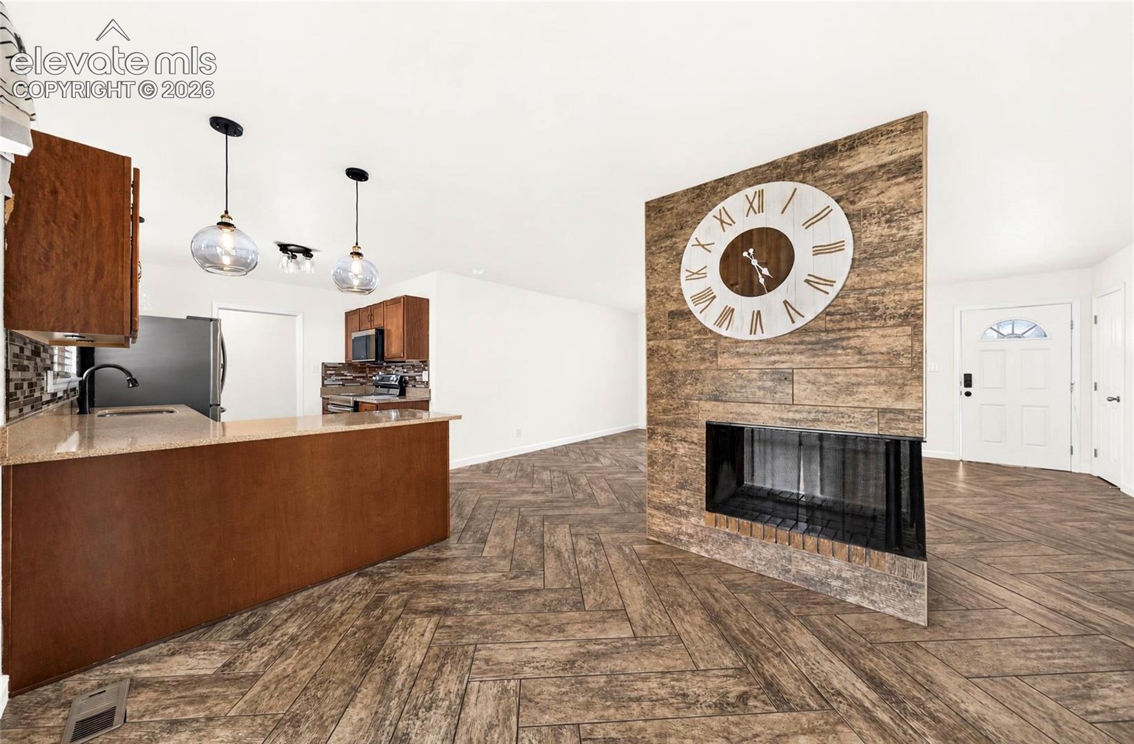 Herringbone pattern wood-finish flooring, two glass pendant lights, stone-finish fireplace surround, and kitchen breakfast bar with stone-finish countertop