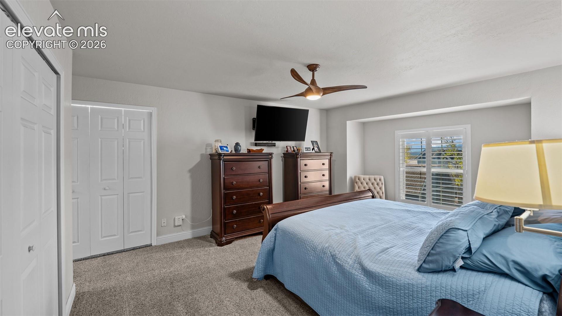 Carpeted room featuring a ceiling fan, dual-panel closet doors, and a window with plantation shutters