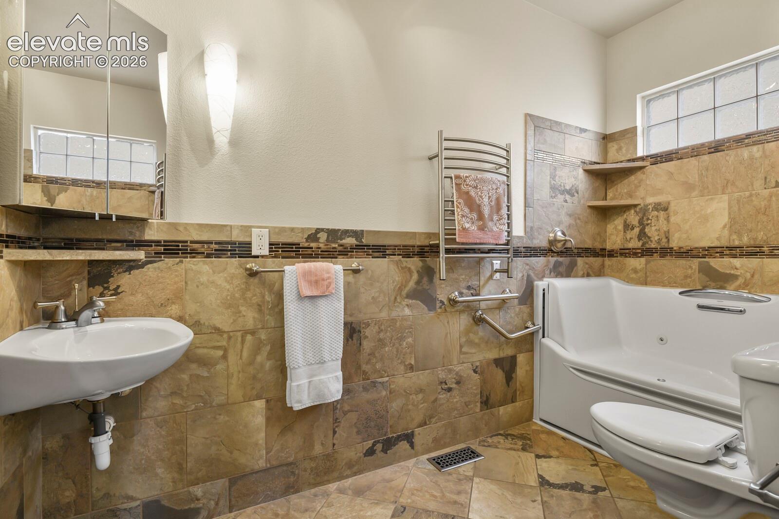 Natural stone wall and floor tiling with decorative glass mosaic accent border, wall-mounted basin, heated towel rack, and walk-in soaking tub with integrated jet features