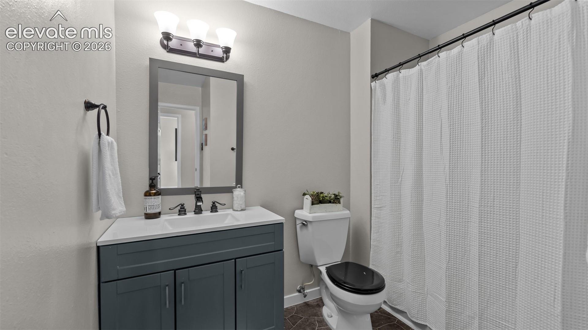Dark-toned vanity with a rectangular sink, stone-composite countertop, matching framed mirror, three-light vanity fixture, and patterned floor tile