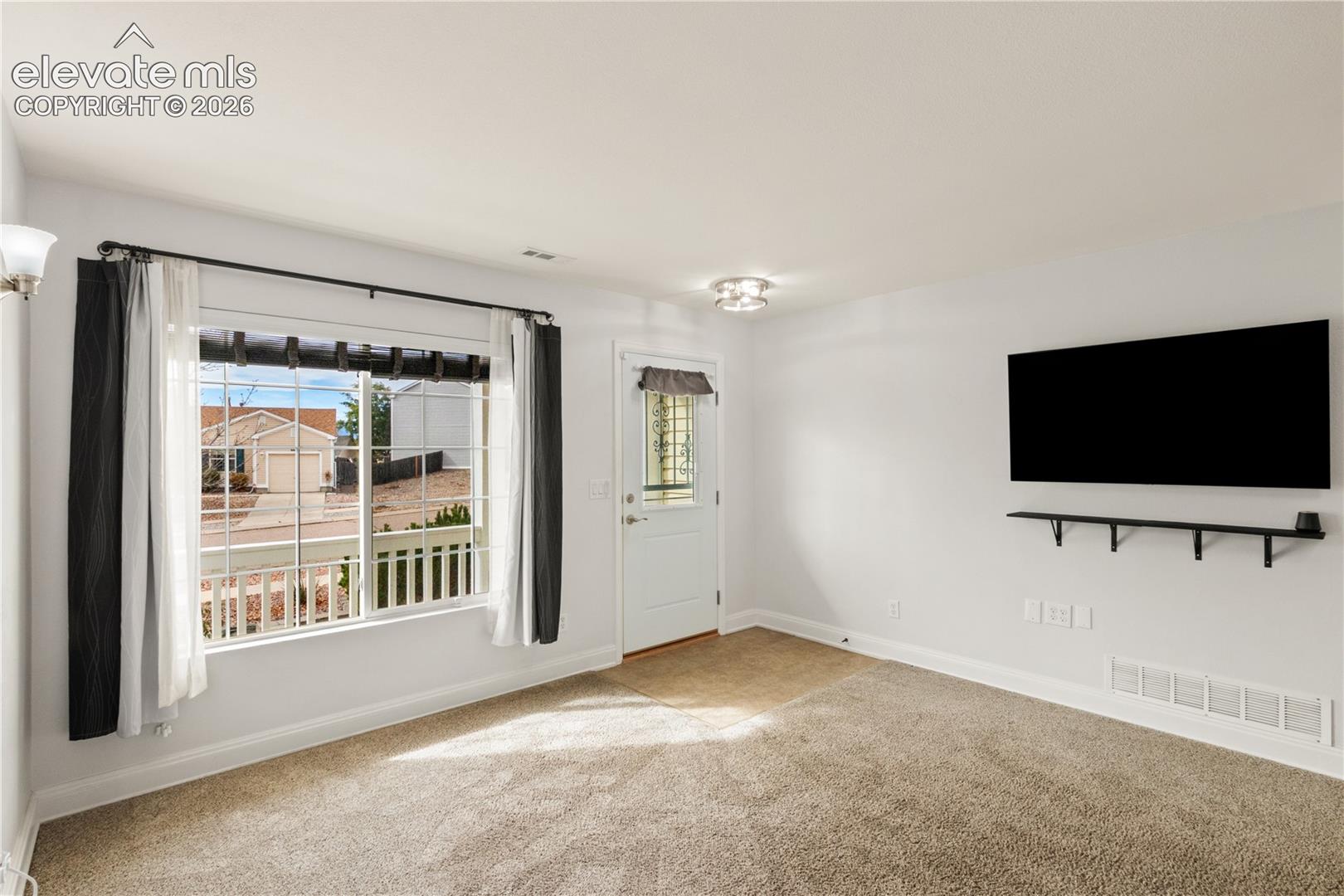 Large window with grid muntins, neutral wall-to-wall carpeting, exterior entry door with integrated window pane, and a wall-mounted floating shelf