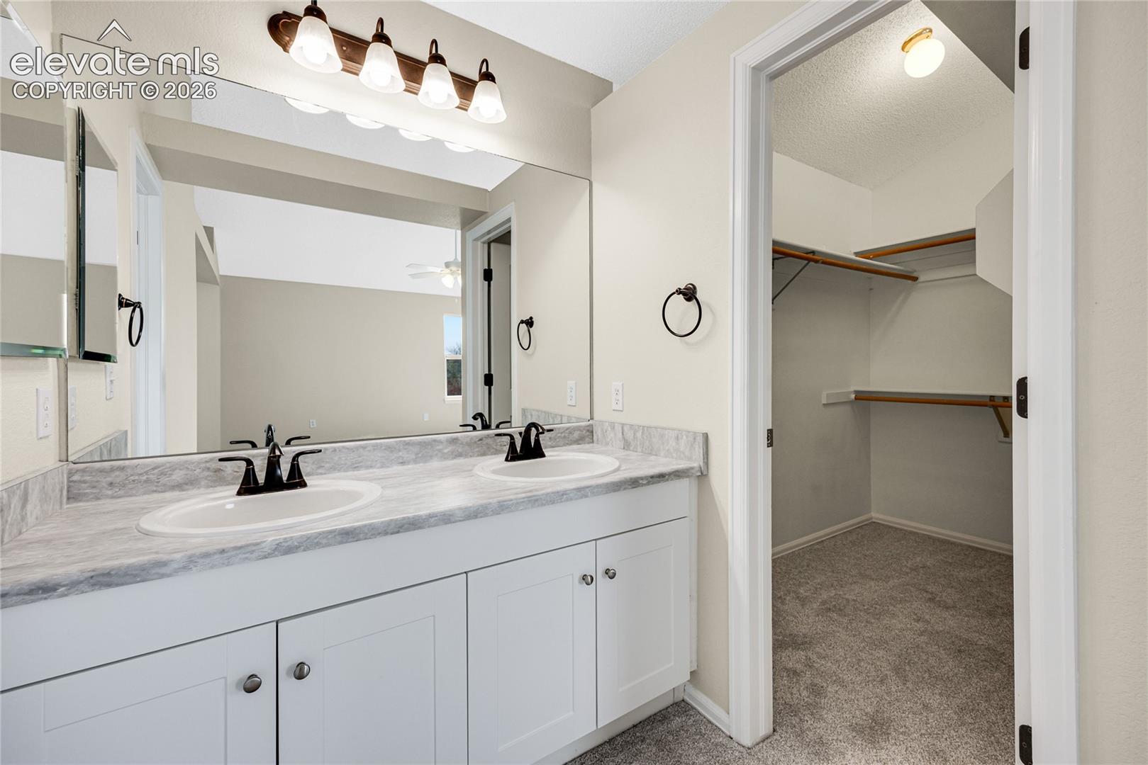 Dual-sink vanity with stone-patterned countertop, white Shaker-style cabinetry, matte black hardware, full-width vanity mirror, and walk-in closet with built-in shelving