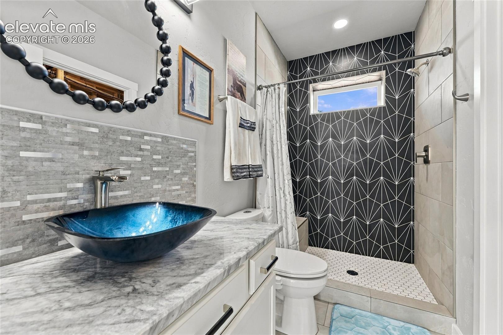 Bathroom featuring a vanity with a stone-finish countertop, blue glass vessel sink, mosaic tile backsplash, and a walk-in shower with geometric accent wall tiling