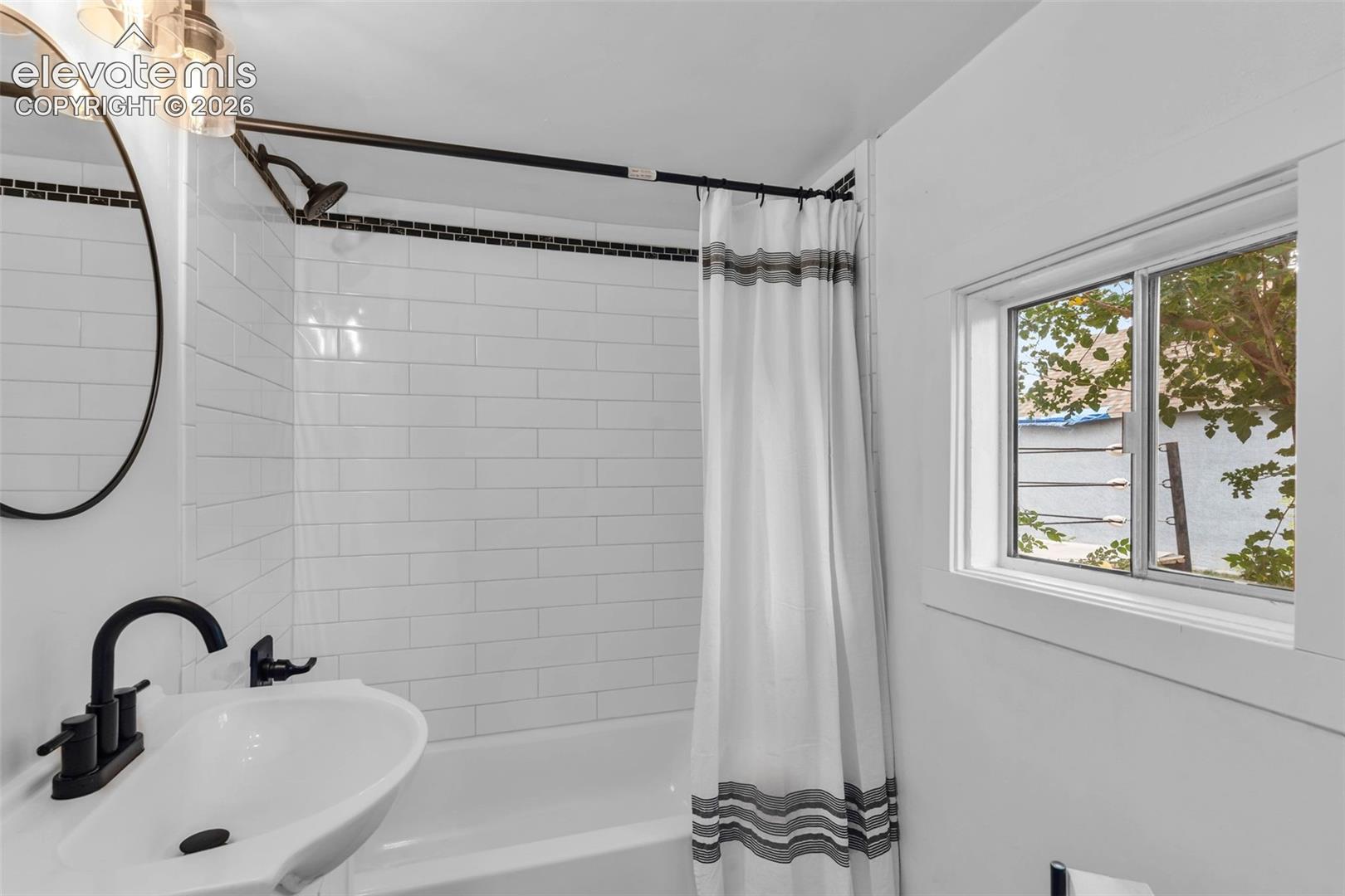 Bathroom featuring white subway tile wall surround with a decorative black tile inlay, matte black wall-mounted faucet, round mirror with a black frame, and a sliding window