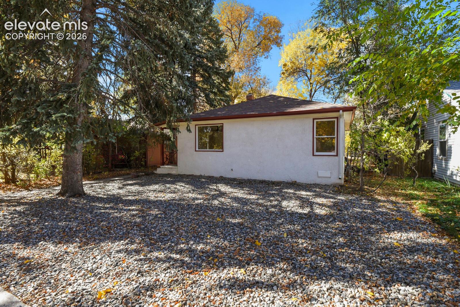 Stucco exterior featuring a hipped roof, stone ground cover, and mature trees