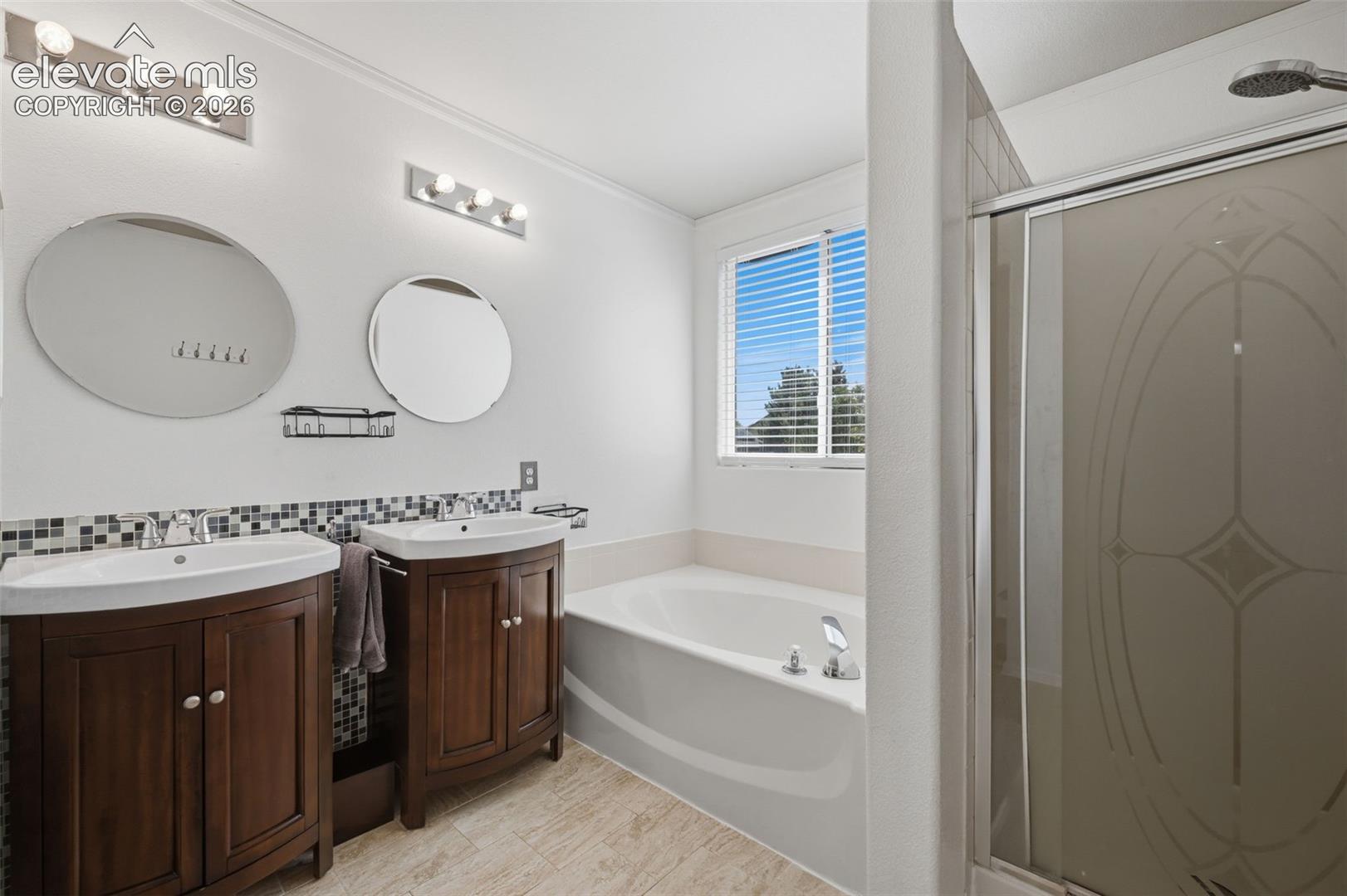 Dual wood-finish vanities with vessel sinks, a jetted soaking tub, tiled flooring, and a glass-enclosed shower with decorative etching