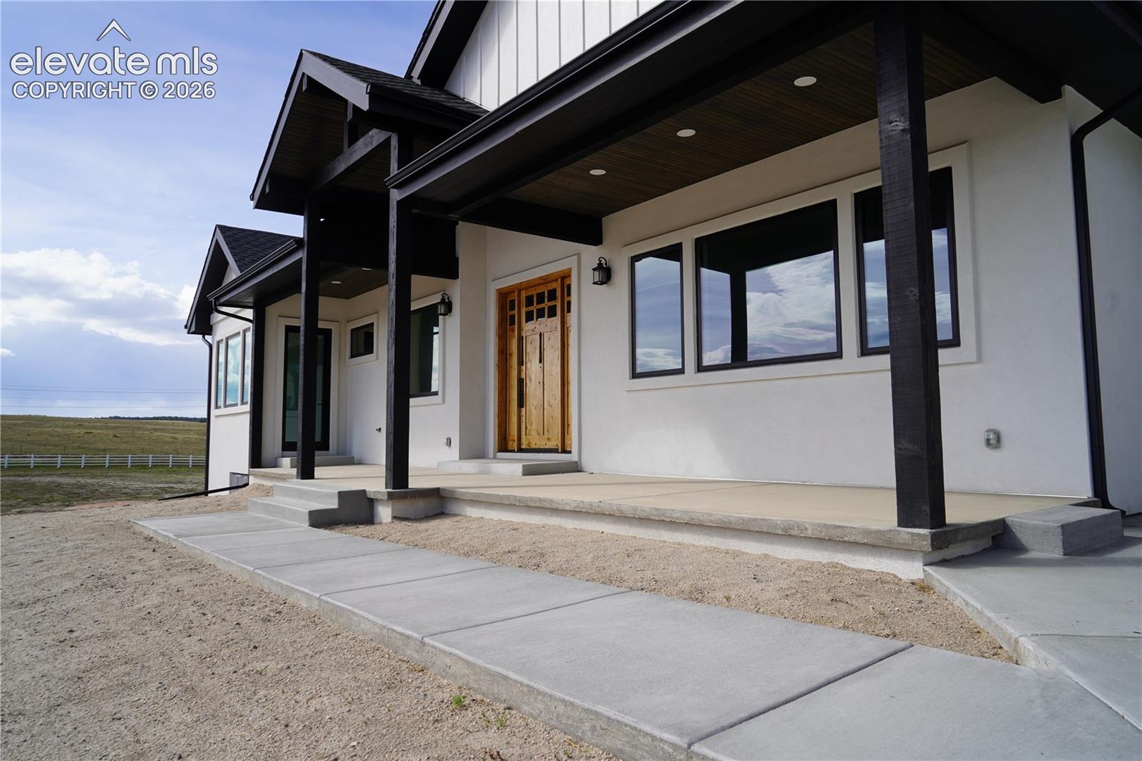 White stucco exterior with black-framed windows, a wood-finish entry door, and a covered porch featuring dark wood columns and a wood-finish ceiling