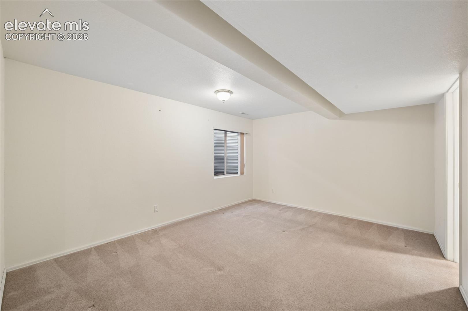 Neutral-toned interior space featuring neutral carpeting, a flush-mount ceiling light fixture, and a window with an egress well