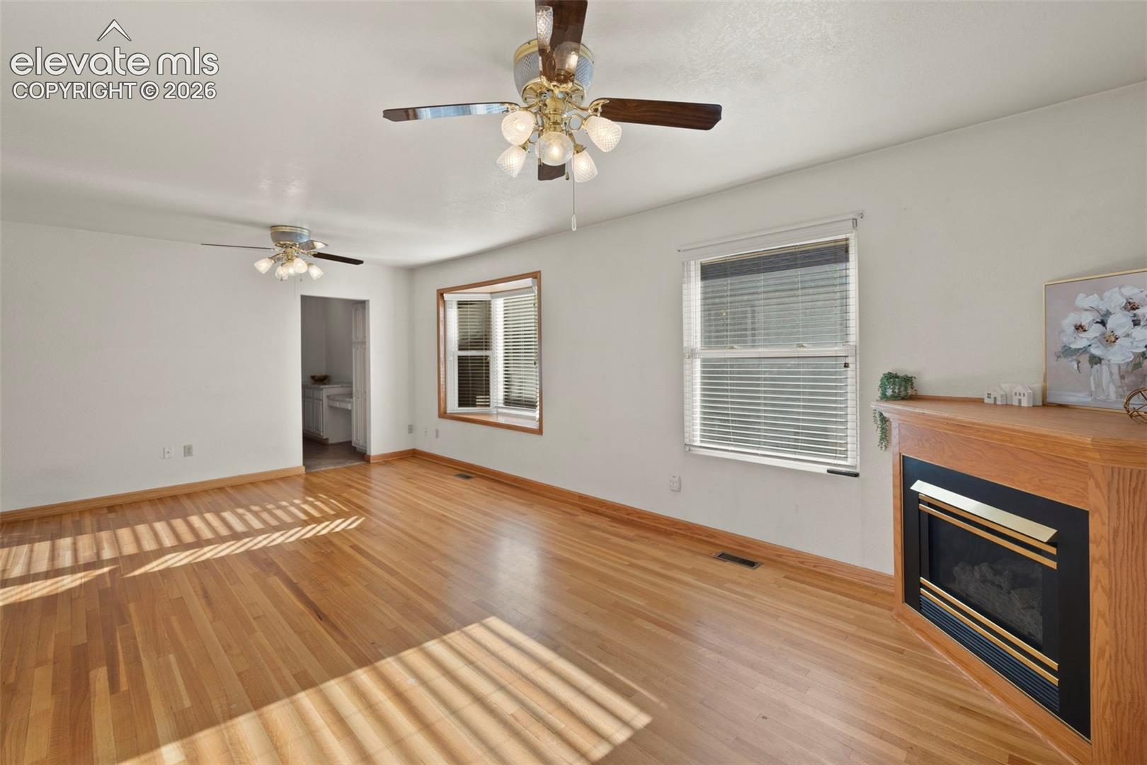 Wood-finish flooring, recessed fireplace with wood surround, multiple windows with blinds, and two ceiling fans with lighting