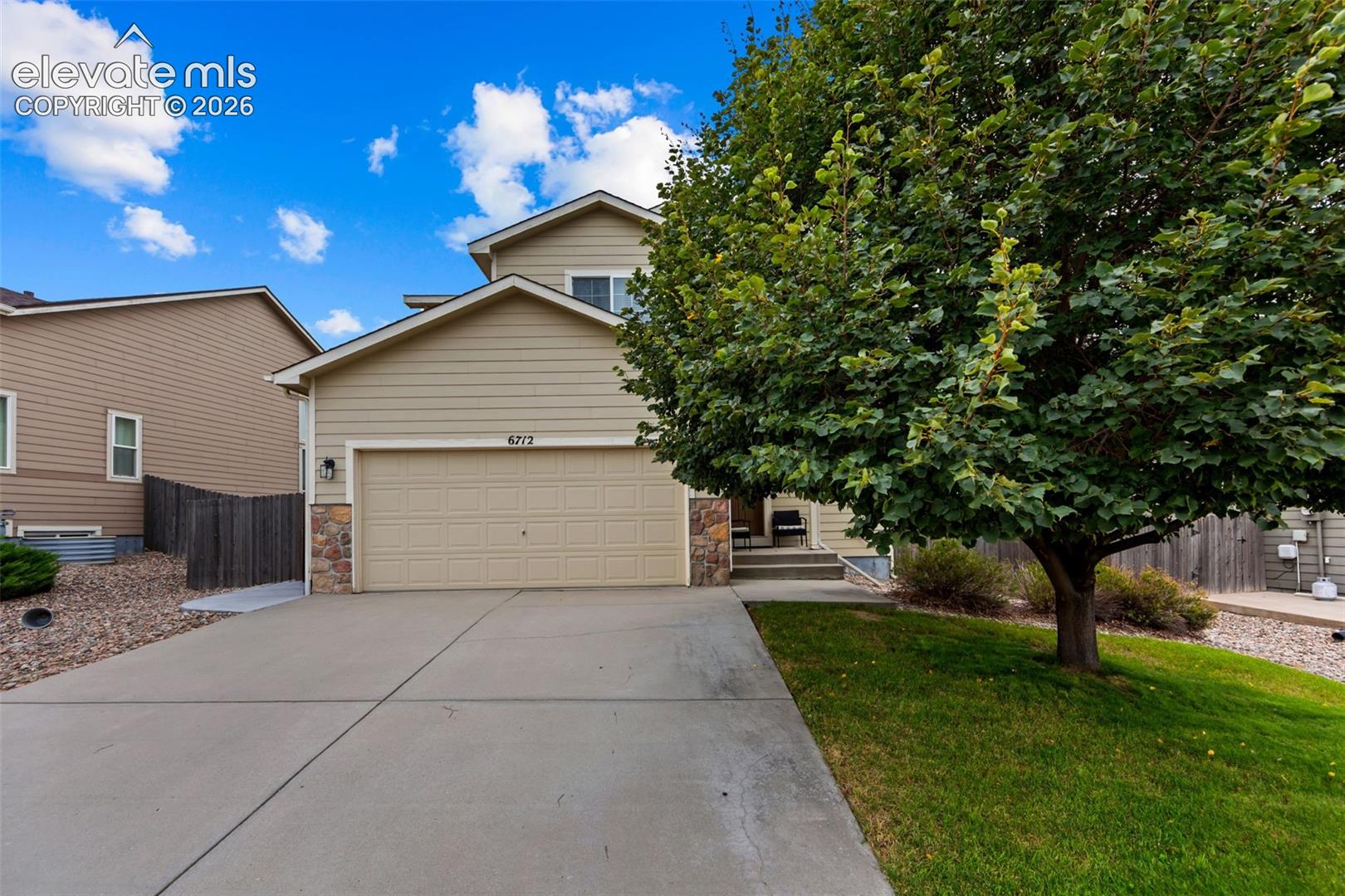 Two-story residence featuring a two-car garage, stone accents, mature front yard tree, and concrete driveway