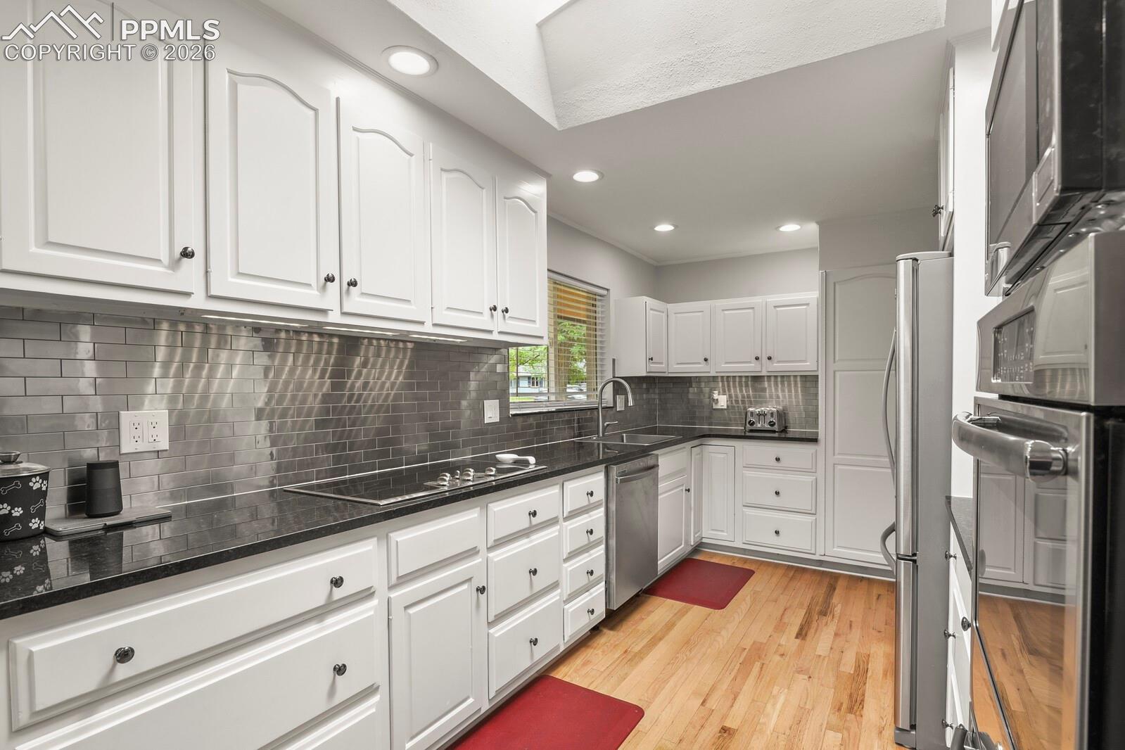 Kitchen featuring white cabinetry, dark stone countertops,  stainless steel tile backsplash, hardwood flooring and recessed lighting