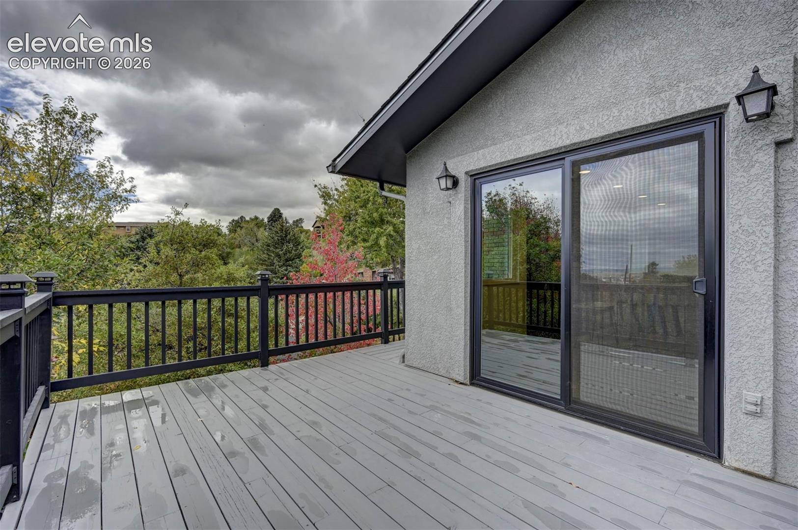 Exterior deck featuring painted wood-finish plank flooring, black metal railing, sliding glass door, and wall-mounted exterior light fixtures