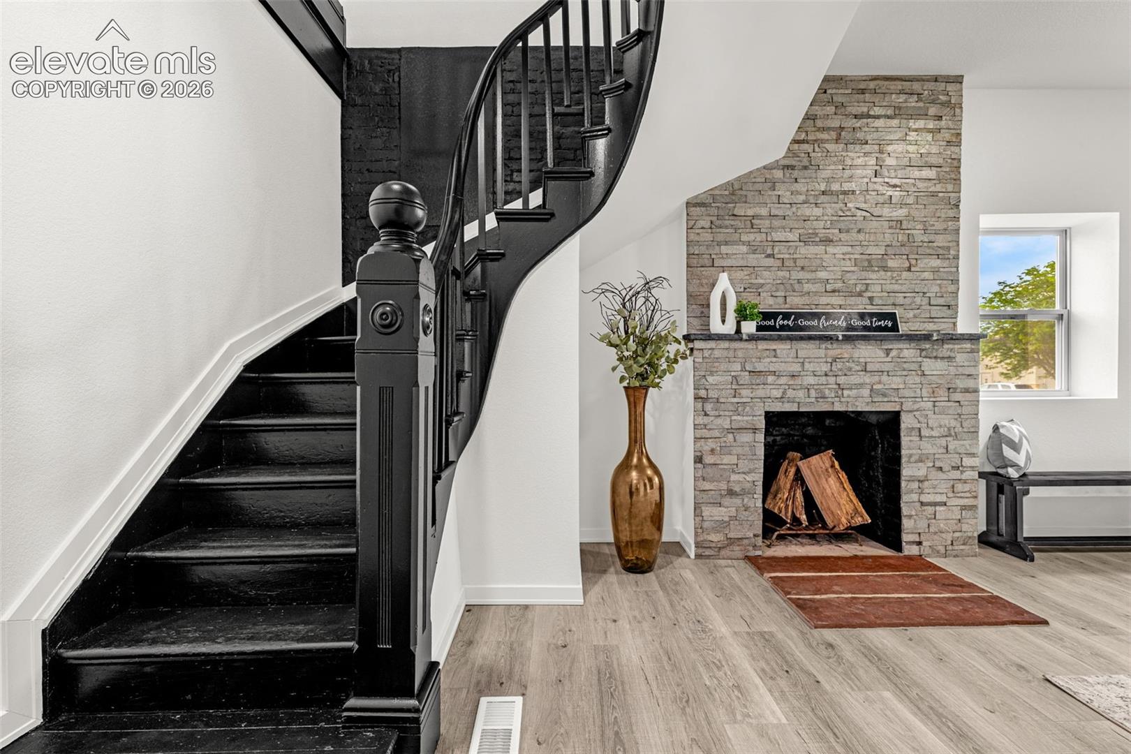 Curved staircase with black newel post and balusters, accompanied by a stacked stone fireplace with a dark mantel, and light wood-finish flooring