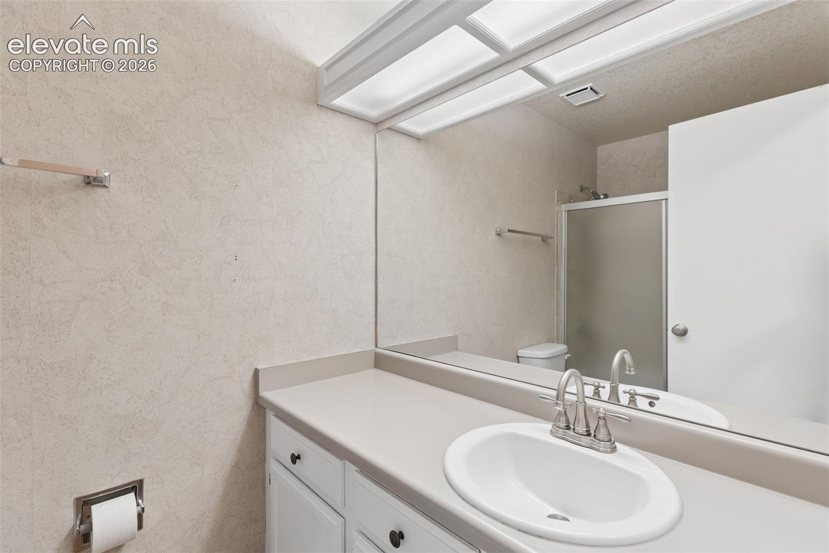 Full-width mirror over a vanity with a solid-surface countertop, white cabinetry, undermount sink with dual-handle faucet, and integrated overhead lighting
