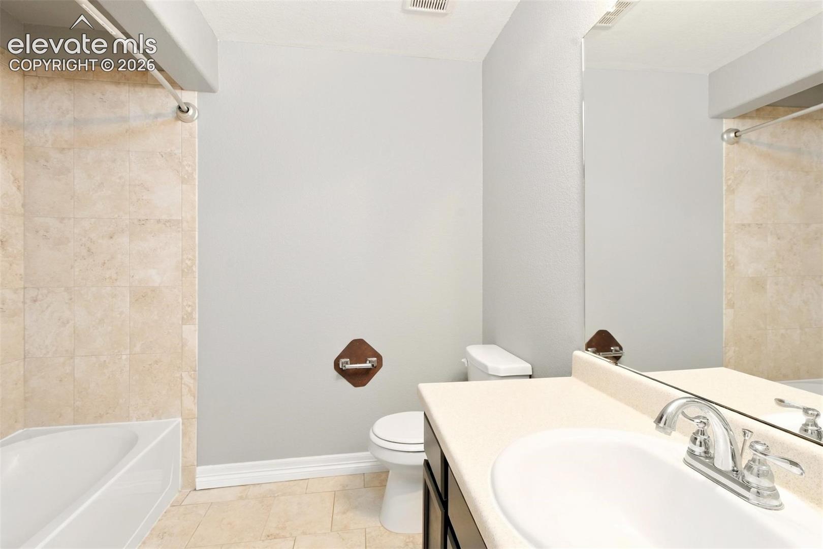 Bathroom featuring a neutral stone-tile tub surround, large wall-mounted mirror, vanity with a solid-surface countertop, and an integrated basin with polished chrome hardware