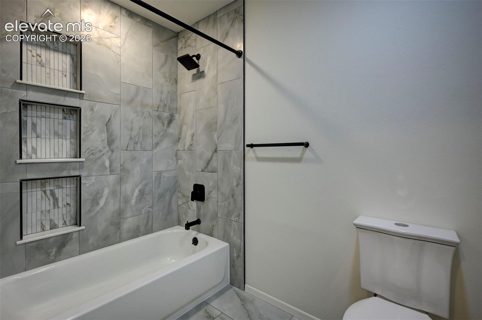 Bathtub featuring large-format stone-look wall tile, triple built-in recessed storage niches, matte black plumbing fixtures, and a coordinated towel bar