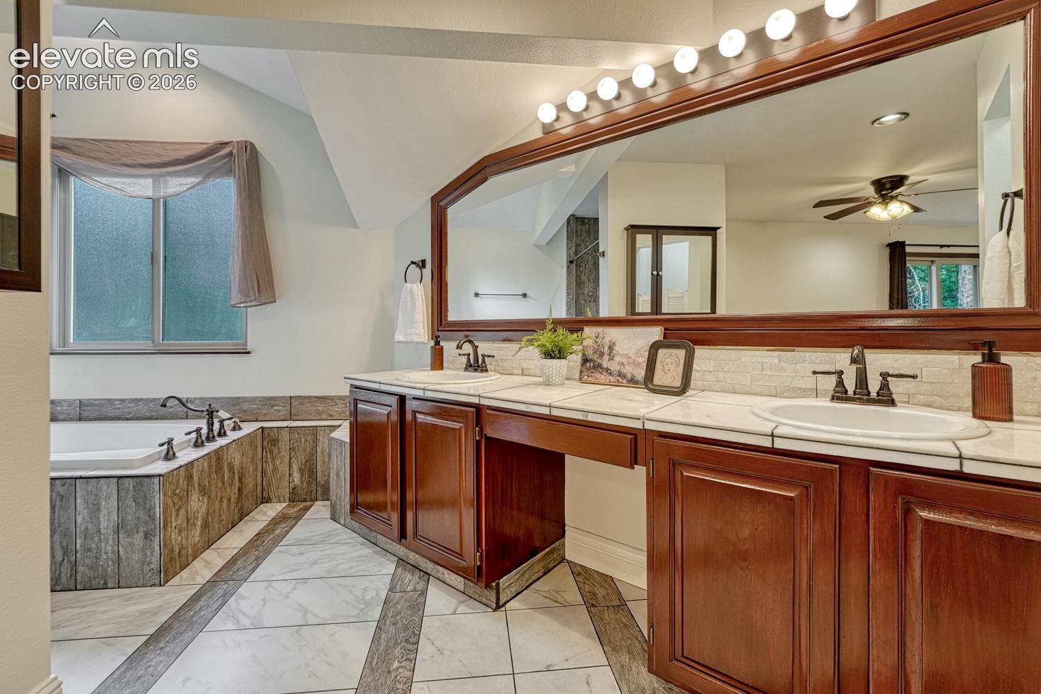 Dual-sink vanity with stained wood cabinetry, tile countertop, large framed wall mirror, multi-light vanity fixture, and tiled soaking tub surround