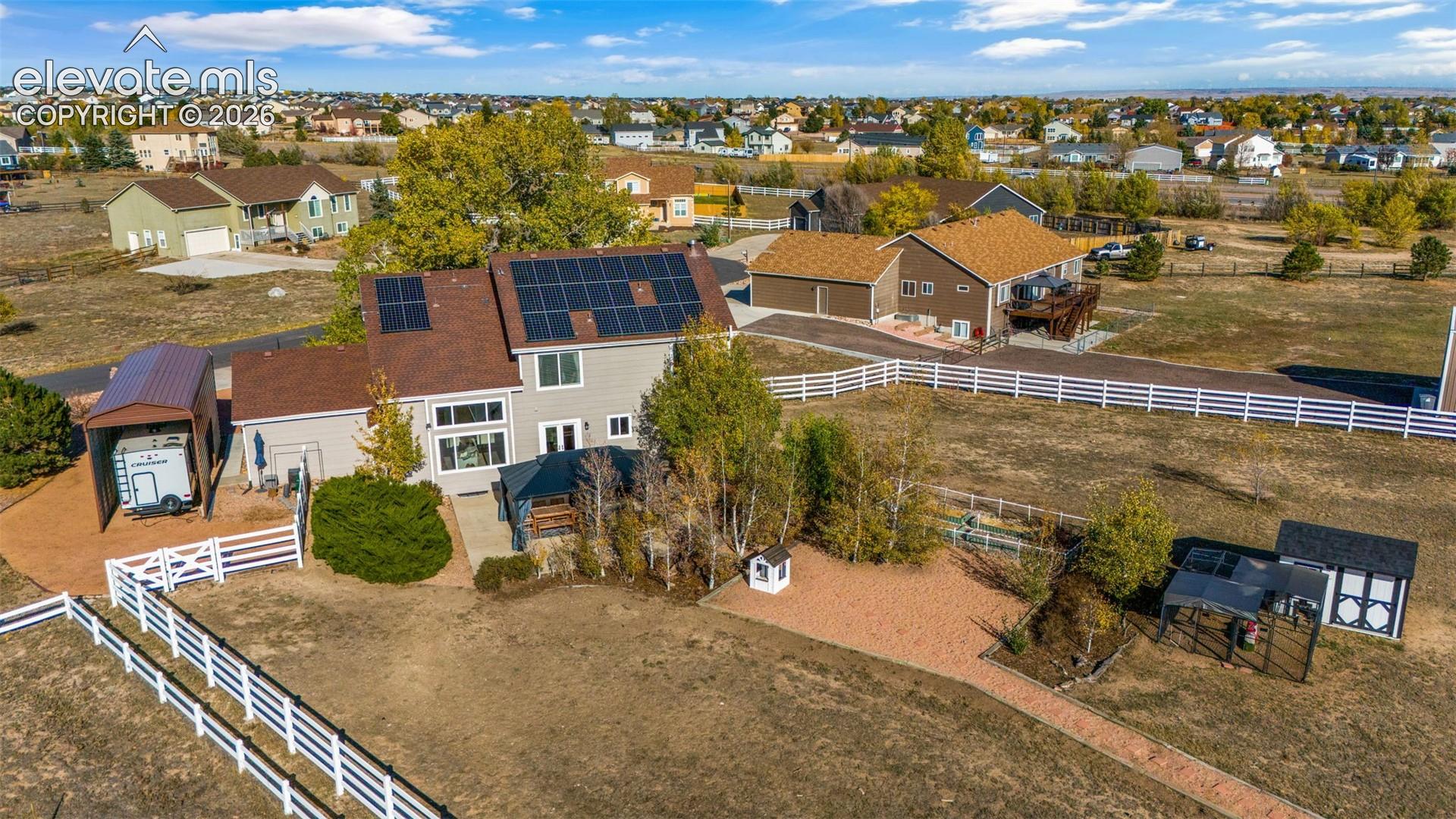 Multi-level residence featuring solar panel array, attached metal-roofed carport, white vinyl post-and-rail perimeter fencing, and detached storage structure