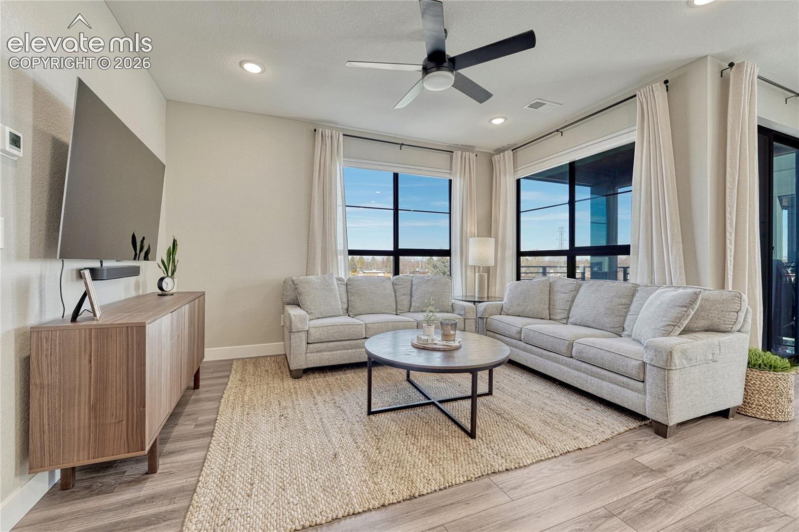 Living area featuring wood-finish flooring, recessed lighting, a modern ceiling fan, and large windows with black frames