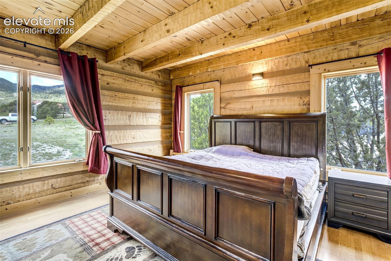 Log cabin interior featuring exposed wood beam ceiling, natural wood walls, light wood-finish flooring, and multiple window openings