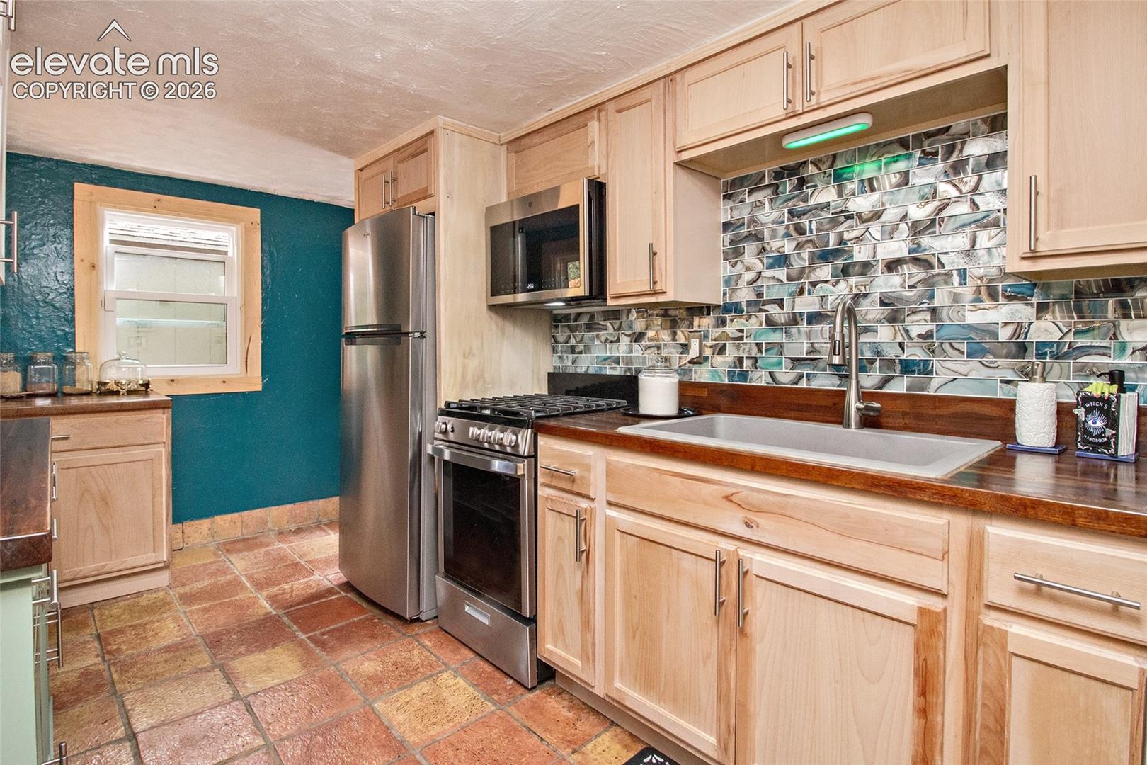 Kitchen featuring natural wood cabinetry, stainless steel appliances, a deep basin sink with a high-arch faucet, a textured tile backsplash, and terra-cotta tile flooring