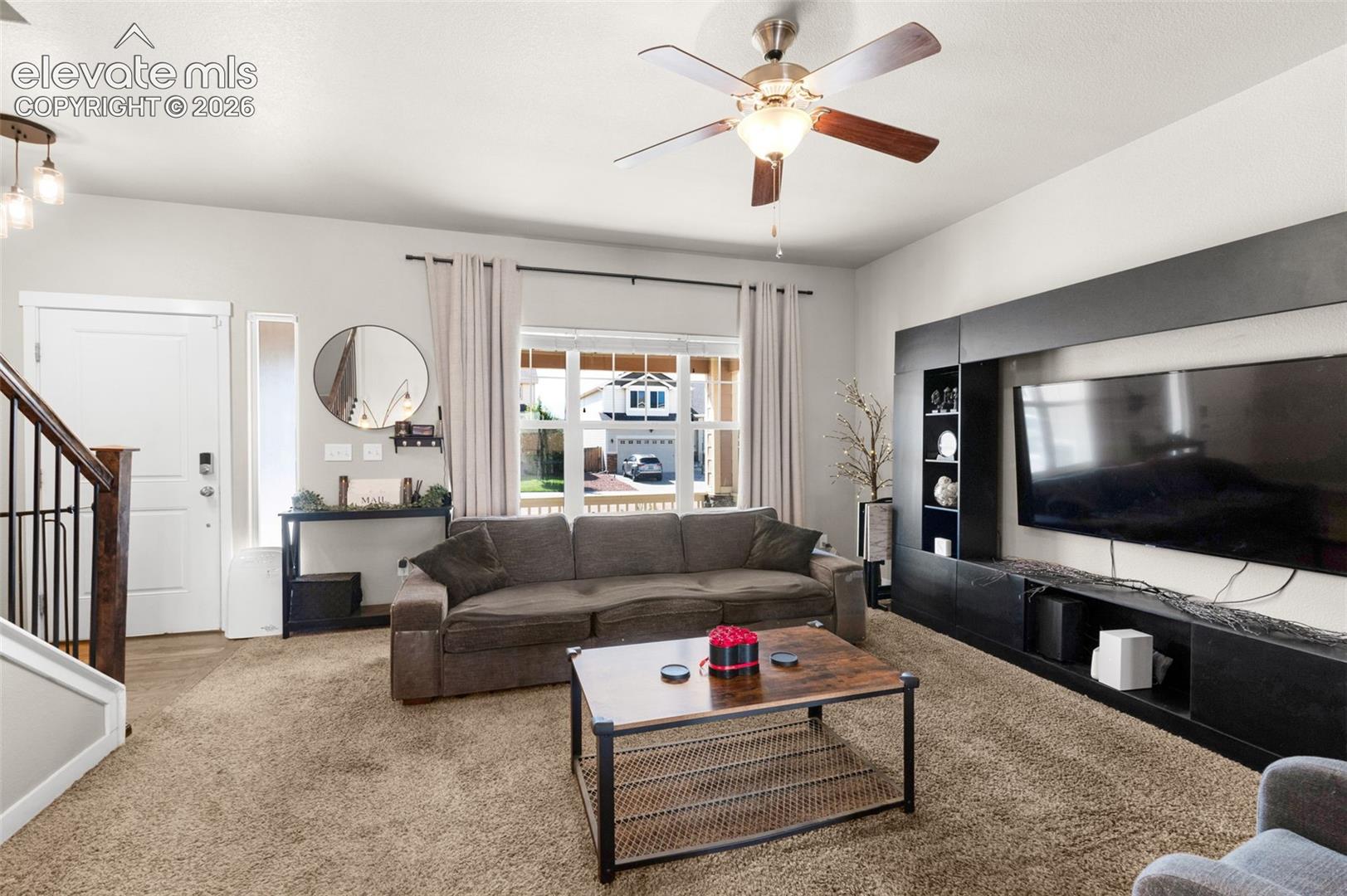Living area featuring wall-to-wall carpeting, ceiling fan with integrated lighting, oversized windows, and built-in black cabinetry with media shelving