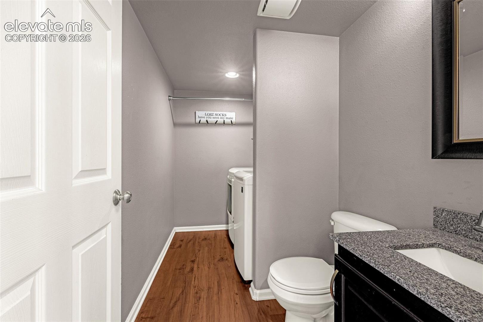 Wood-finish flooring, vanity with stone-composite countertop, white toilet, recessed lighting, and wall-mounted garment rod