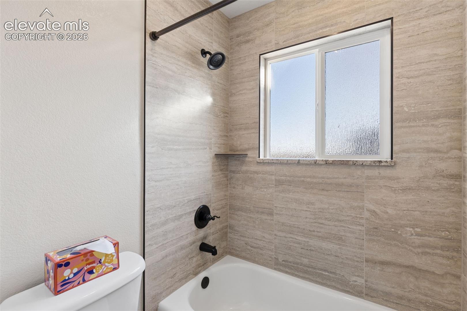 Full-height stone-finish tile shower surround, matte black rainfall showerhead and faucet fixtures, and an integrated recessed shelf
